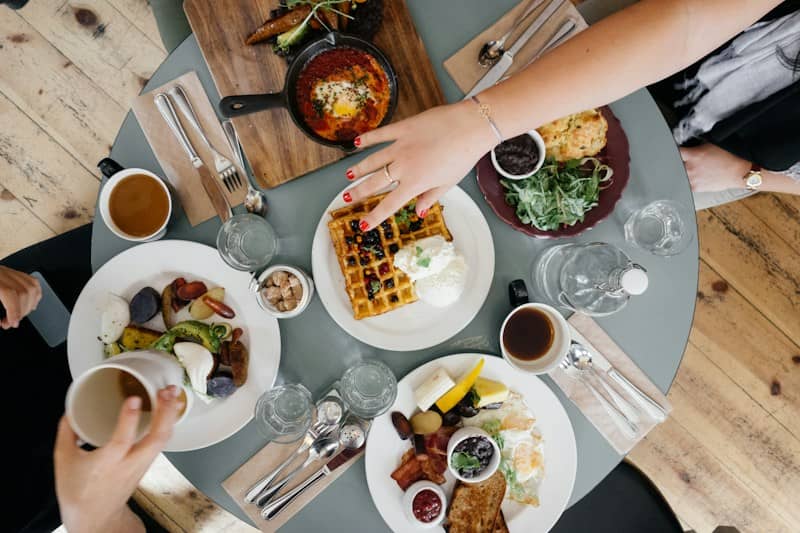 Hands reaching across a table. The table is full of plates and pans and glasses and utensils. One hand has red nailpolish, a ring, and a bracelet. There is a second, unadorned hand. The table is grey.