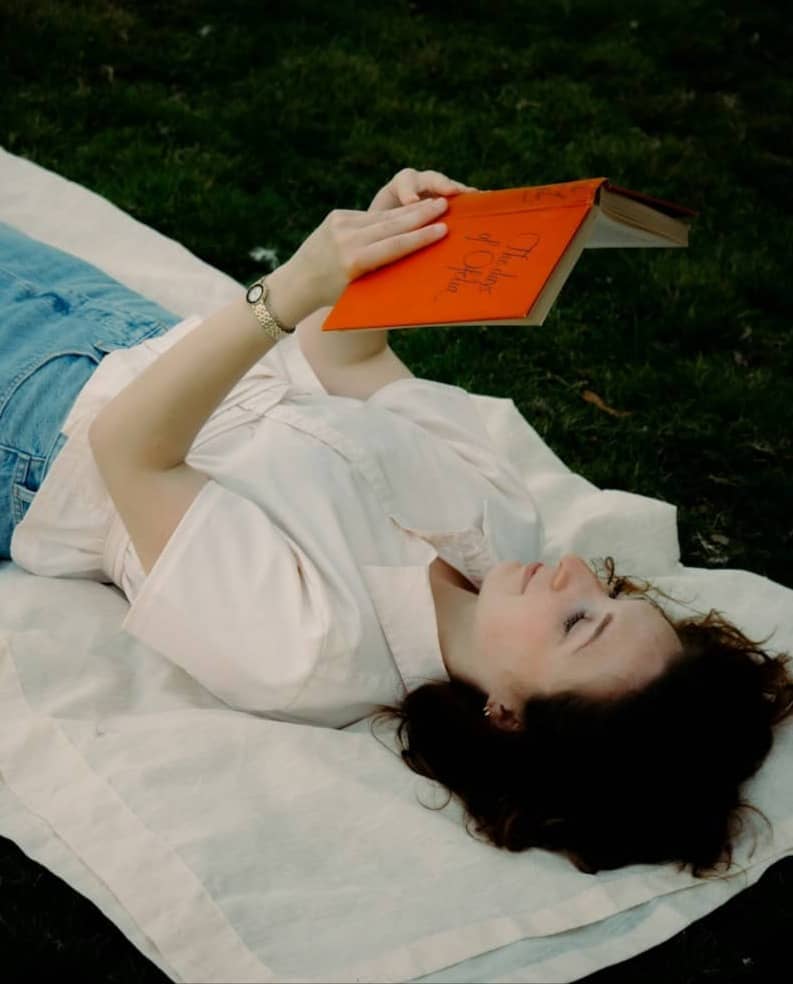 A woman in a white shirt reading an orange book while lying on a blanket on grass