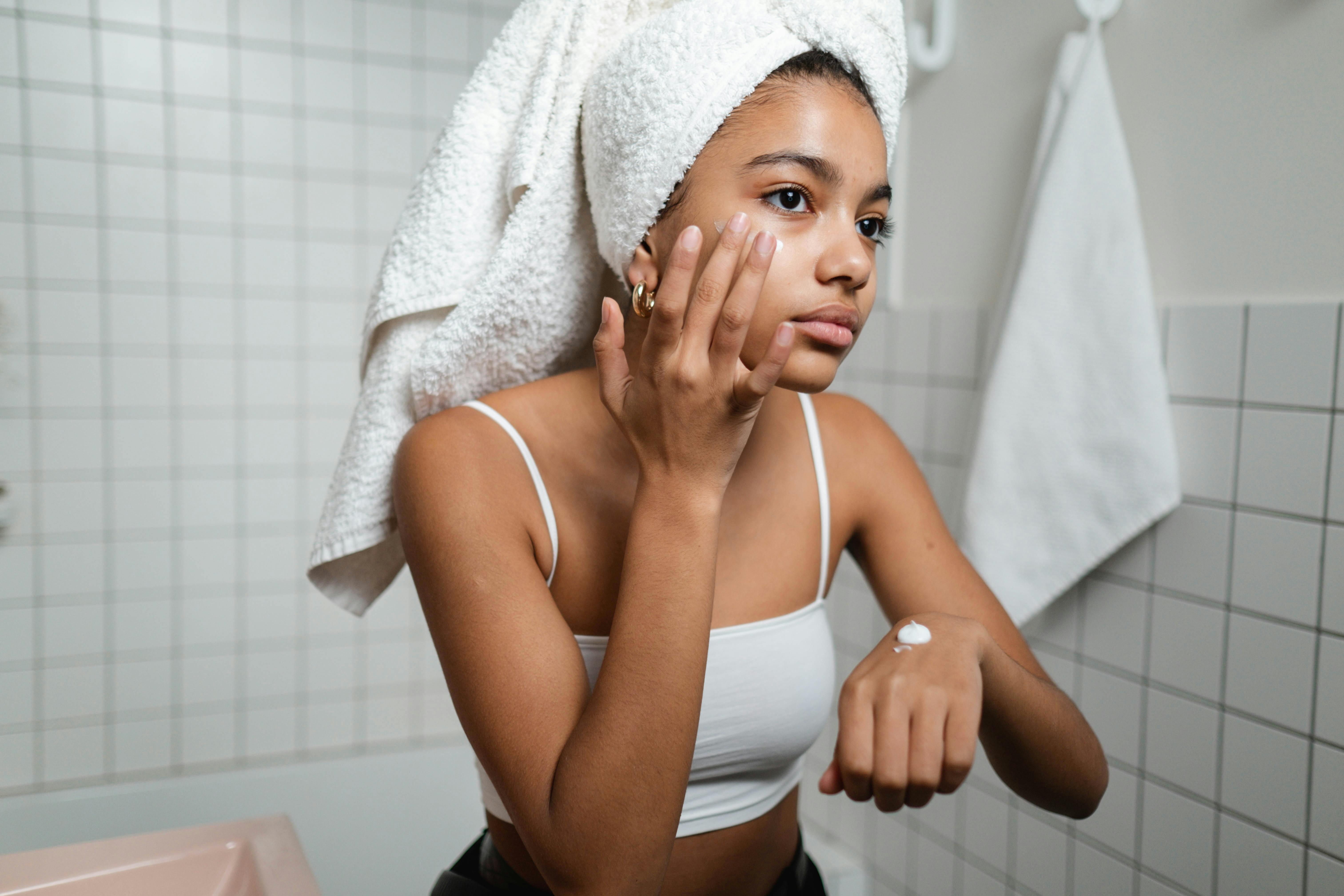 A person applying cream to their face, focusing on the cheek area. The individual has a gentle expression and is wearing a textured headband. The image is a close-up, highlighting smooth skin and well-manicured nails.