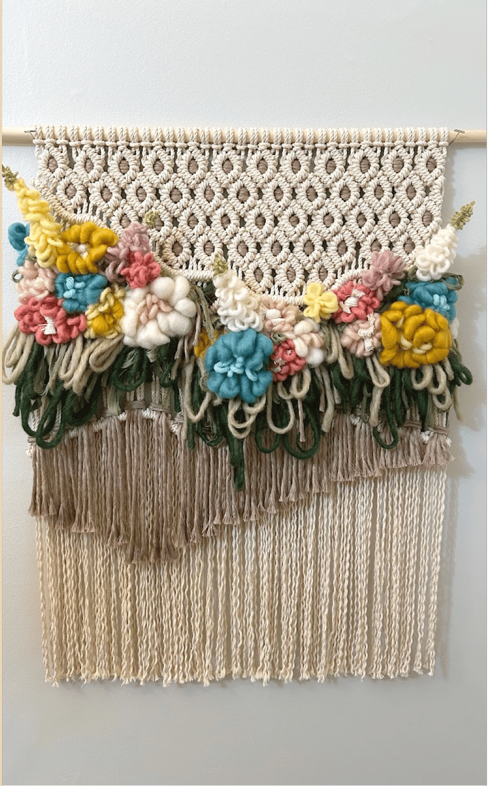 A handmade macramé wall hanging featuring intricate patterns and colorful yarn flowers. The top part has a diamond pattern in cream-colored yarn, with an array of vibrant flowers in shades of yellow, blue, pink, and white mixed with greenery. Below, long strands of neutral-colored yarn hang down.