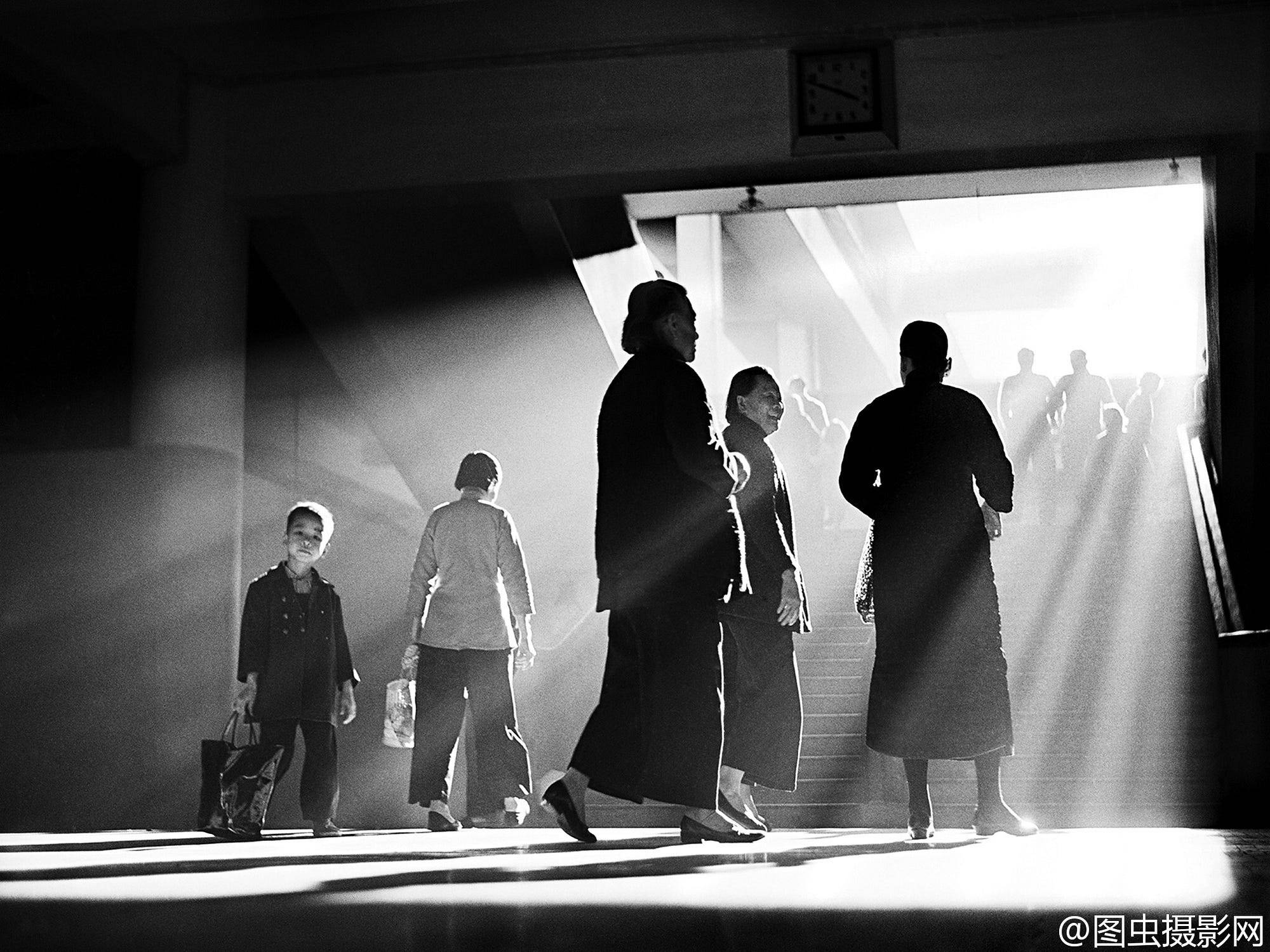     Black-and-white street photograph of people walking through a bright stairwell as sunlight streams across the floor and silhouettes their figures.