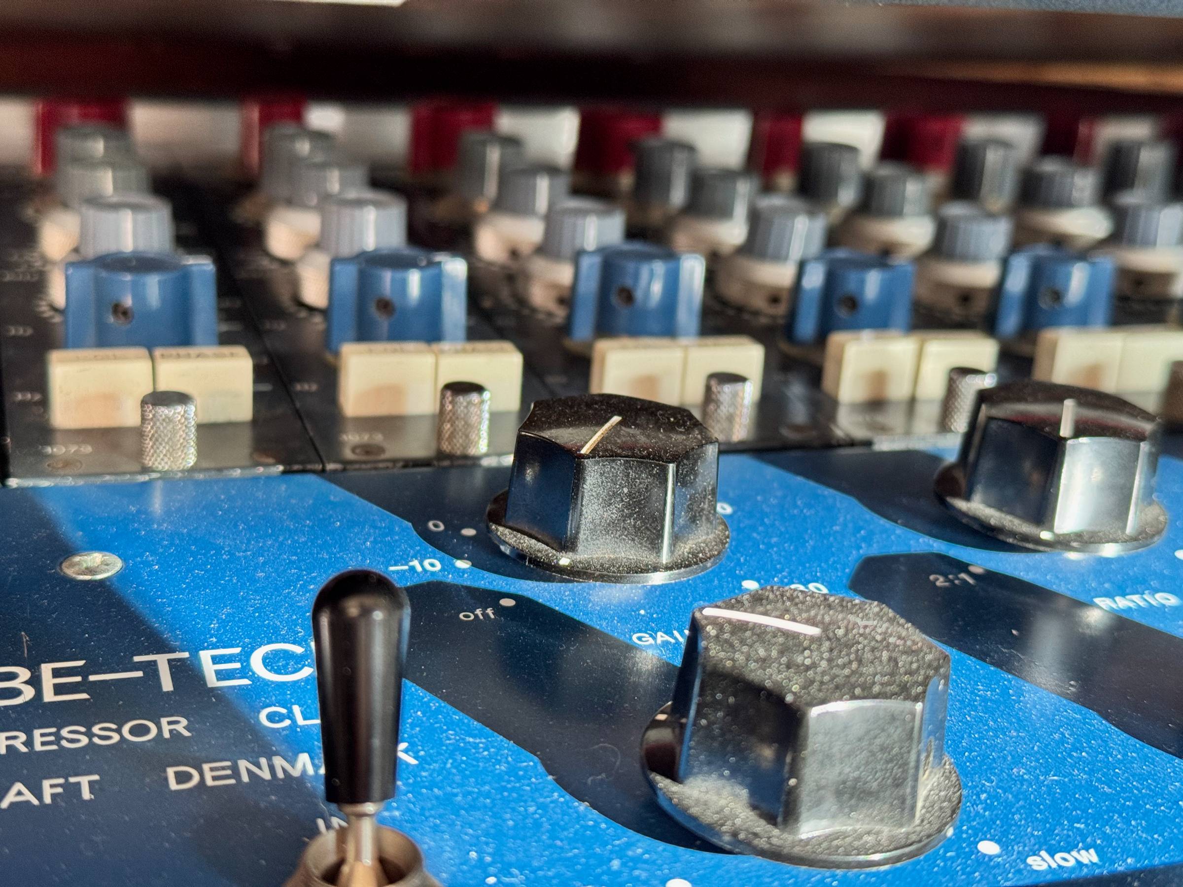     Close-up of blue analog audio compressor hardware with black control knobs, toggle switch, and blurred mixing console controls behind it.