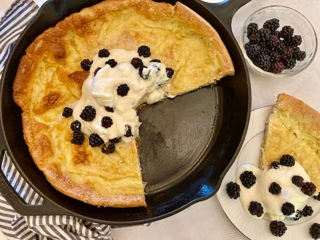 Oat Dutch baby topped with whipped cream and blackberries in a cast iron skillet with a slice on a plate.