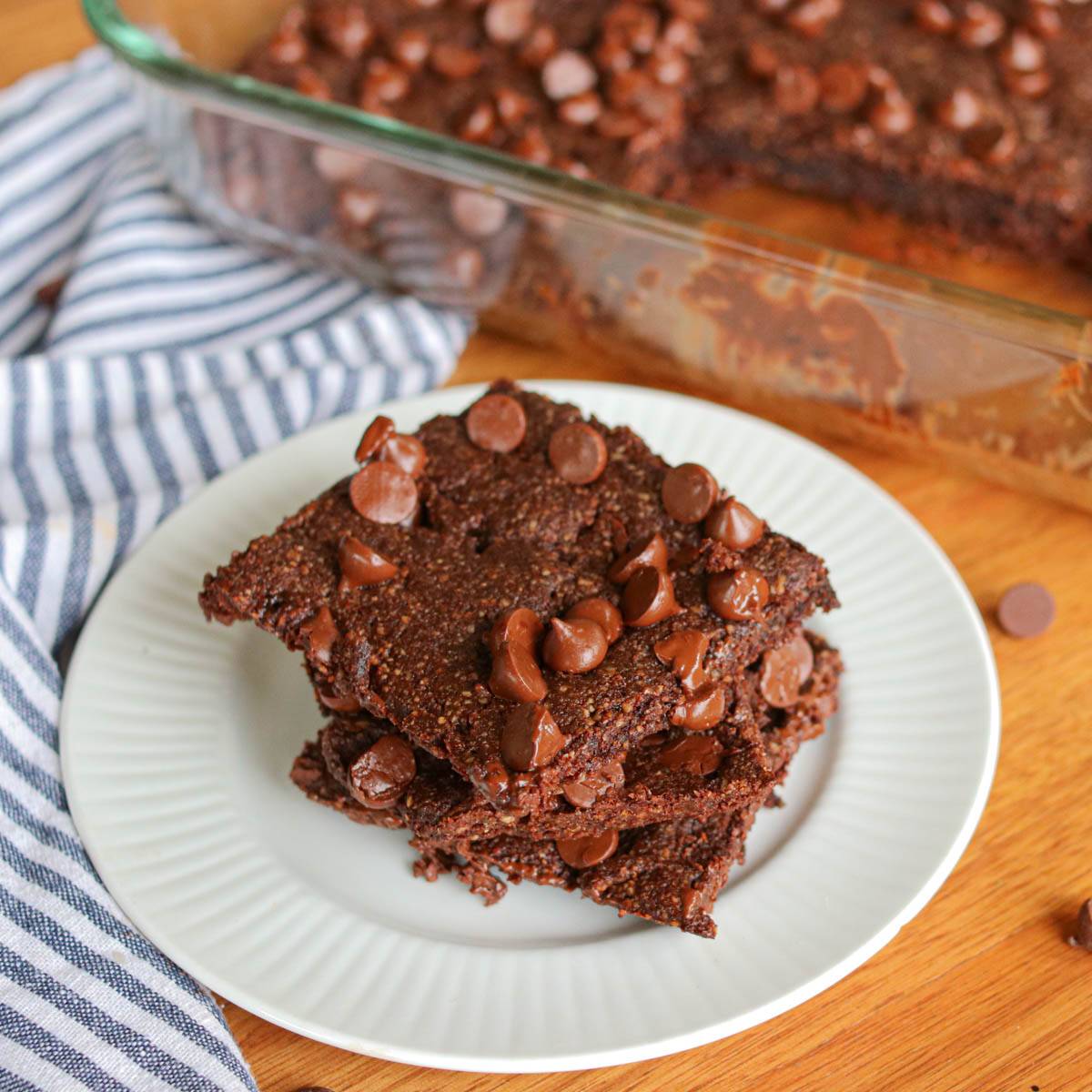 Stack of 3 almond flour brownies on a white plate with a pan of brownies in the background.