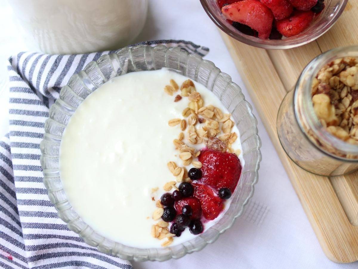 A bowl of homemade yogurt topped with granola and berries with a striped napkin and more toppings in the background.