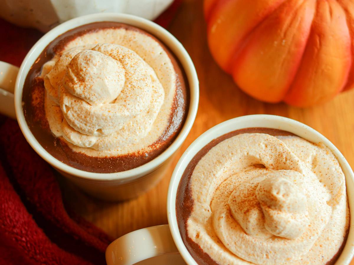 Two cups with pumpkin spice hot chocolate topped with whipped cream on a wooden table.