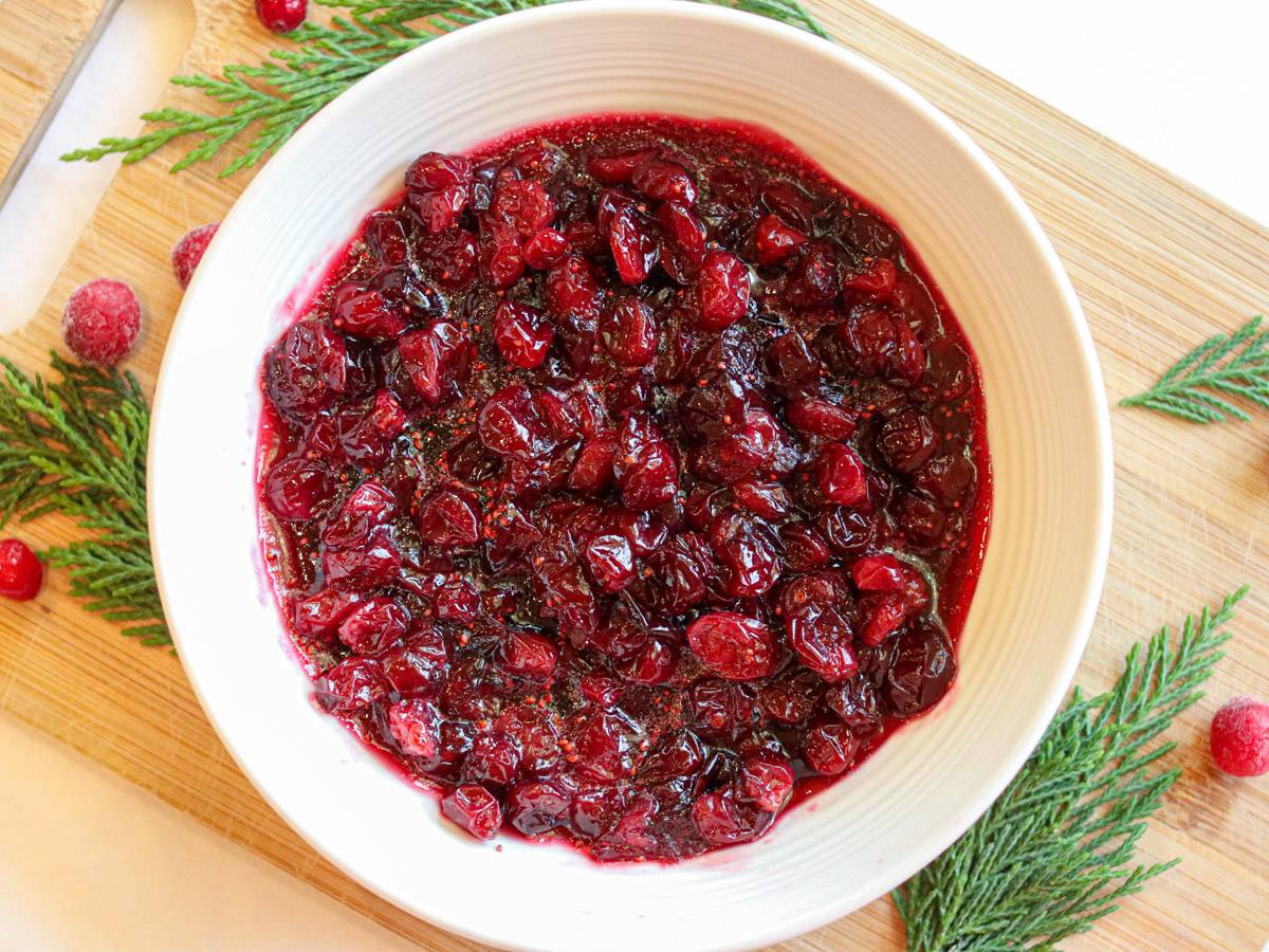 Healthy 3-ingredient cranberry sauce in a bowl on a cutting board surrounded by evergreen leaves and frozen cranberries.