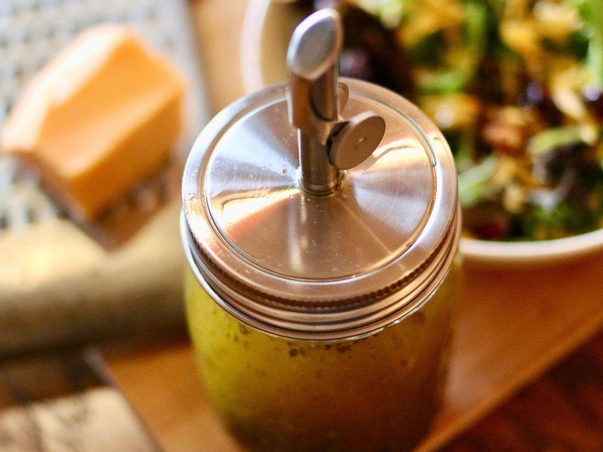 A jar of homemade Italian vinaigrette dressing beside a salad and block of cheese on a grater.