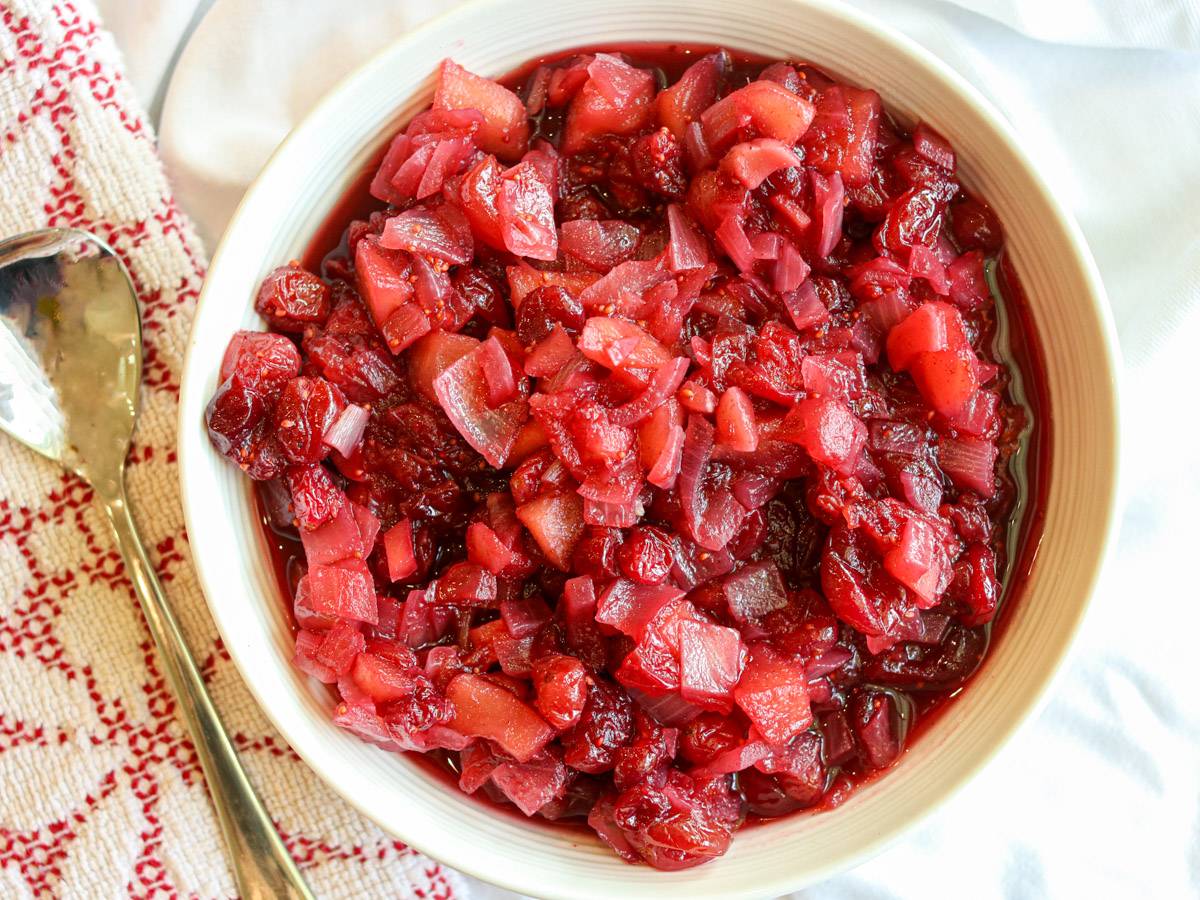 Apple cranberry chutney in a white bowl beside a red and white kitchen towel and serving spoon. 