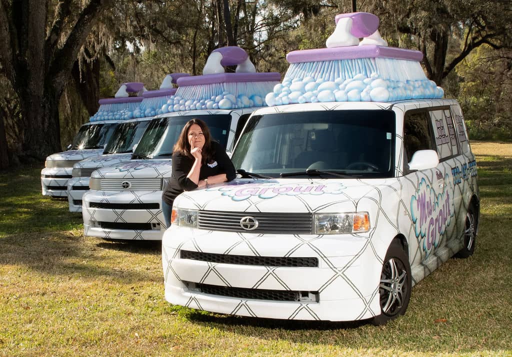 Lisa, owner of Mrs. Grout, pictured with 4 "Grout-mobiles." Vehicles are decorated with a kitchen tile design and have giant scrub brushes with bubbles on the roofs.
