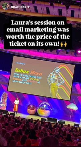Laura Belgray stands on stage presenting to a seated audience in a large auditorium. The stage is decorated with oversized, colorful props including balloons and clouds. A large screen behind her displays a slide titled "Inbox Hero" with a photo of the presenter, Laura Belgray, in a striped top and yellow pants. The slide text reads: "How to write emails that have your list clicking, buying, and hanging on your every word." At the top of the image, white text on a black background reads: "Laura’s session on email marketing was worth the price of the ticket on its own! 🙌" The Instagram handle @upwriteltd is in the top left corner.