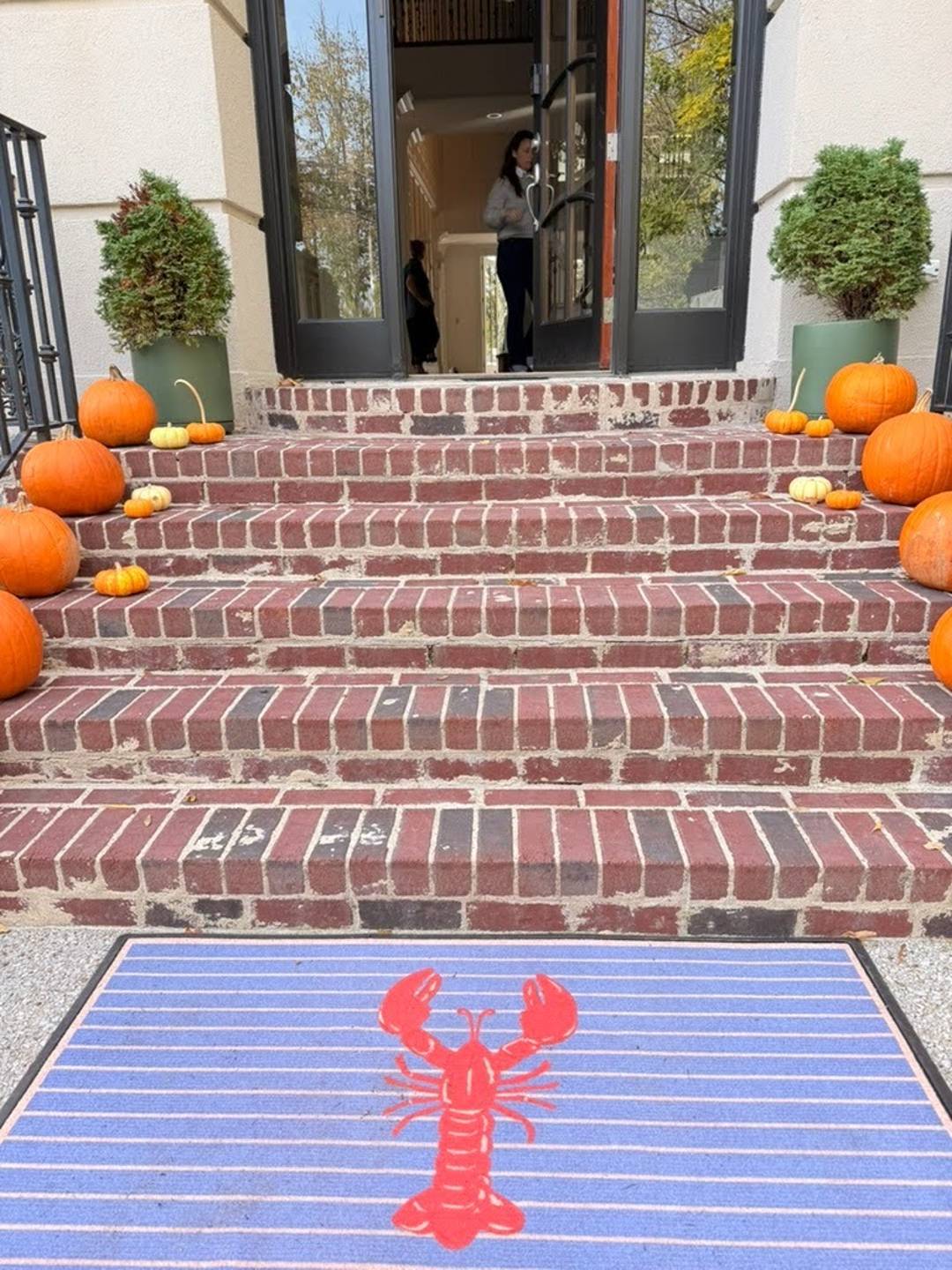 Blue striped doormat with a red lobster at the base of brick front steps lined with orange pumpkins. Potted evergreen shrubs flank an open doorway with two people visible inside.
