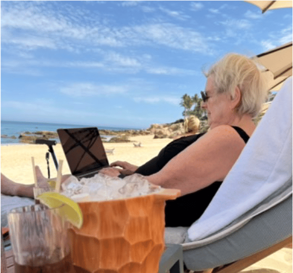 An older woman with short light hair sits in a beach lounge chair using a laptop while facing the ocean. She is under a beach umbrella with sandy shoreline, rocks, and blue water in the background. In the foreground, a wooden ice bucket filled with ice and a drink with a lime wedge sit on a nearby table.