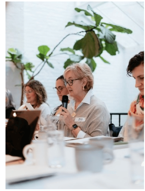 Woman with short blonde hair and glasses speaking into a handheld microphone while seated at a table during a workshop, with other participants listening and taking notes in a bright room with large plants in the background.