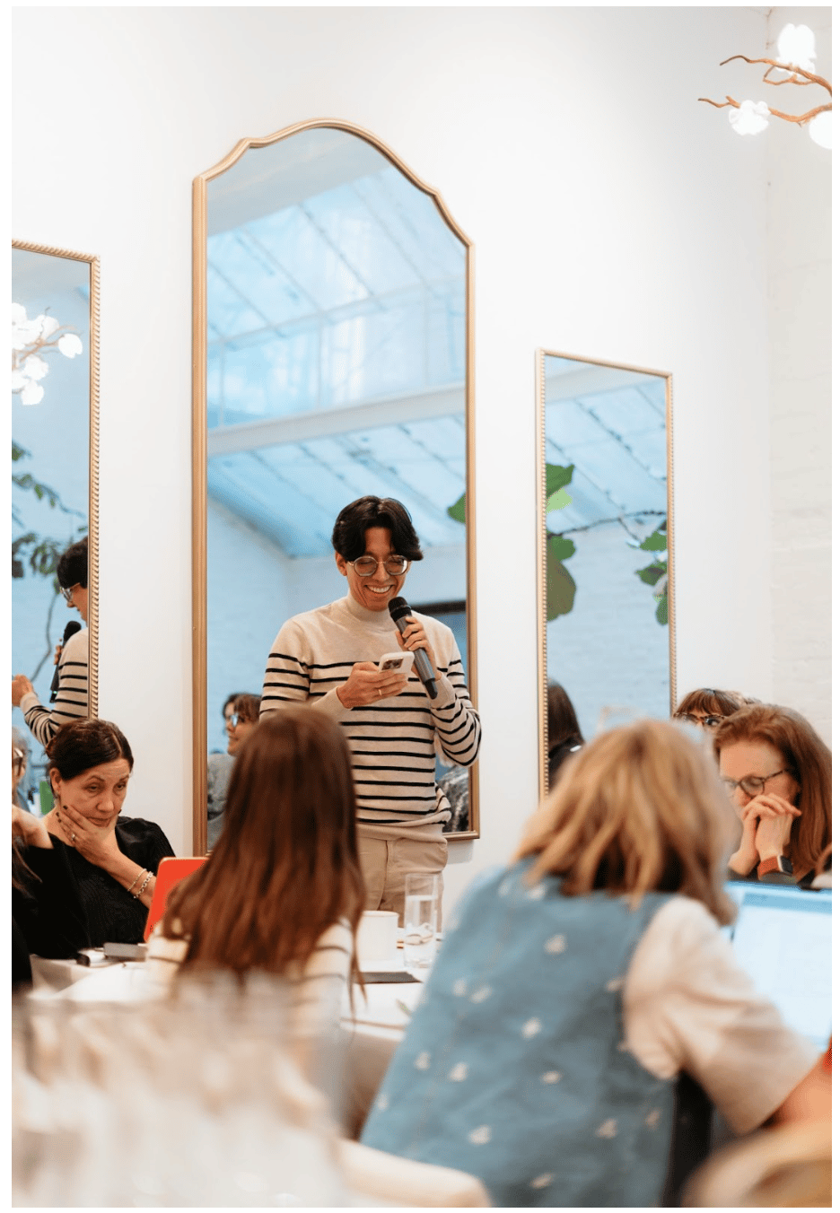 Person standing and smiling while reading from a phone into a handheld microphone during a workshop, with attendees seated around tables listening and working in a bright room with large mirrors and plants.