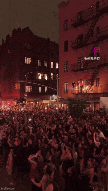 A gif of a nighttime street celebration shows a large crowd packed into an intersection, cheering, dancing, and jumping with their hands in the air. Apartment buildings surround the scene, with people gathered on fire escapes and leaning out of windows to watch. Many attendees are holding up phones to record the moment, while the crowd below appears to be celebrating enthusiastically together. 