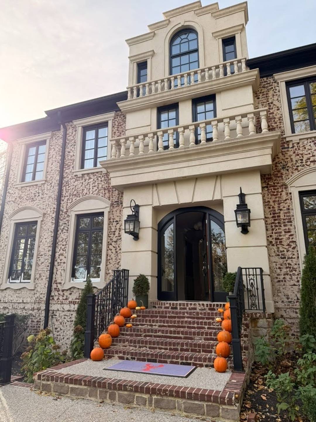 Orange pumpkins line brick steps leading to an arched black front door on a brick house with cream stone trim, decorative balconies, black iron railings, and lantern-style lights.