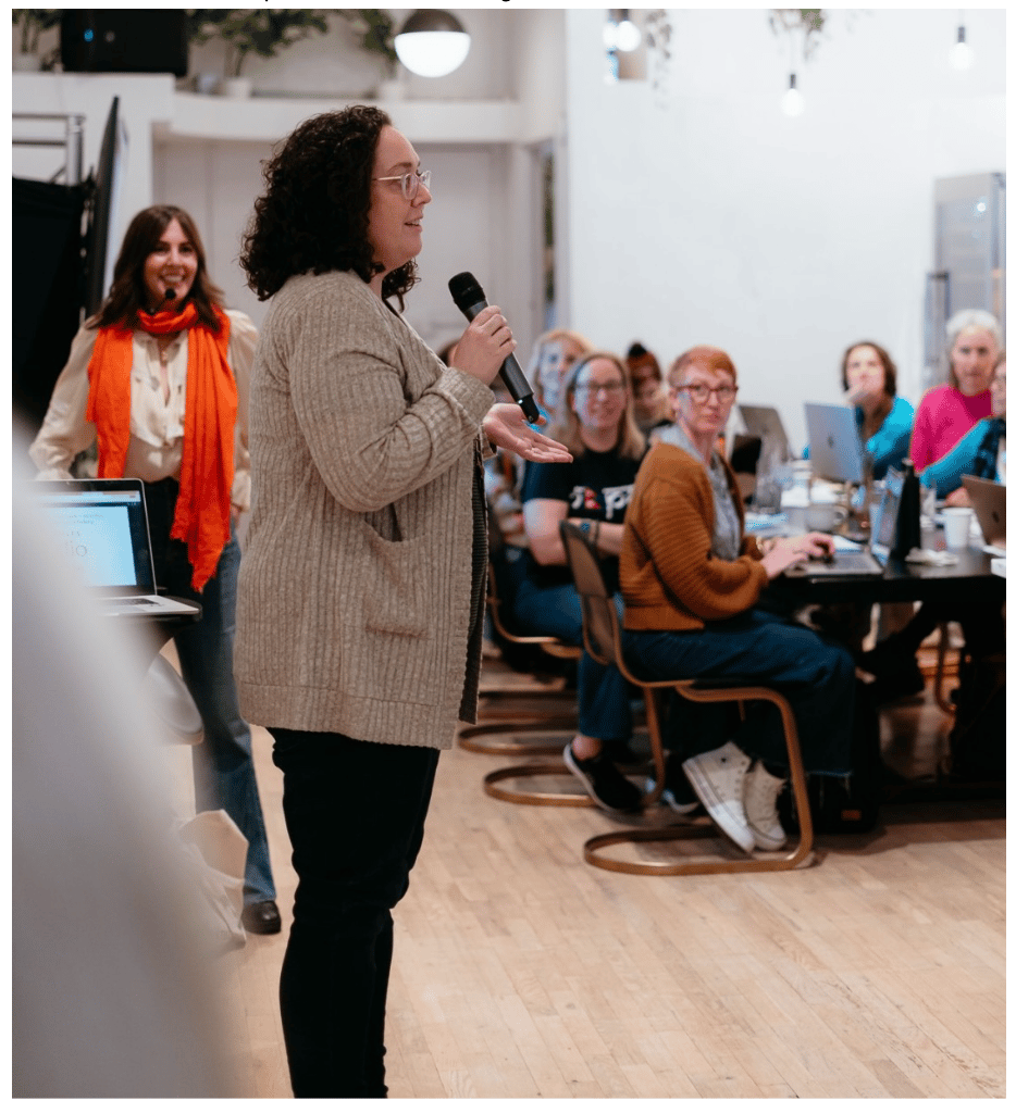 Woman with curly hair and glasses speaking into a handheld microphone during a workshop, standing in front of a seated audience working on laptops in a bright, modern room, while another woman with an orange scarf smiles in the background.