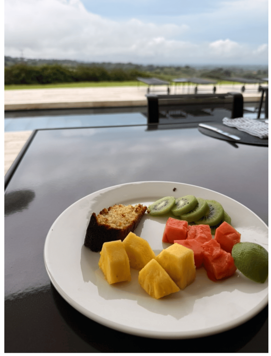 Plate with sliced kiwi, papaya, pineapple, a piece of cake, and a lime wedge on a glass table overlooking an outdoor pool and distant greenery under a cloudy sky.