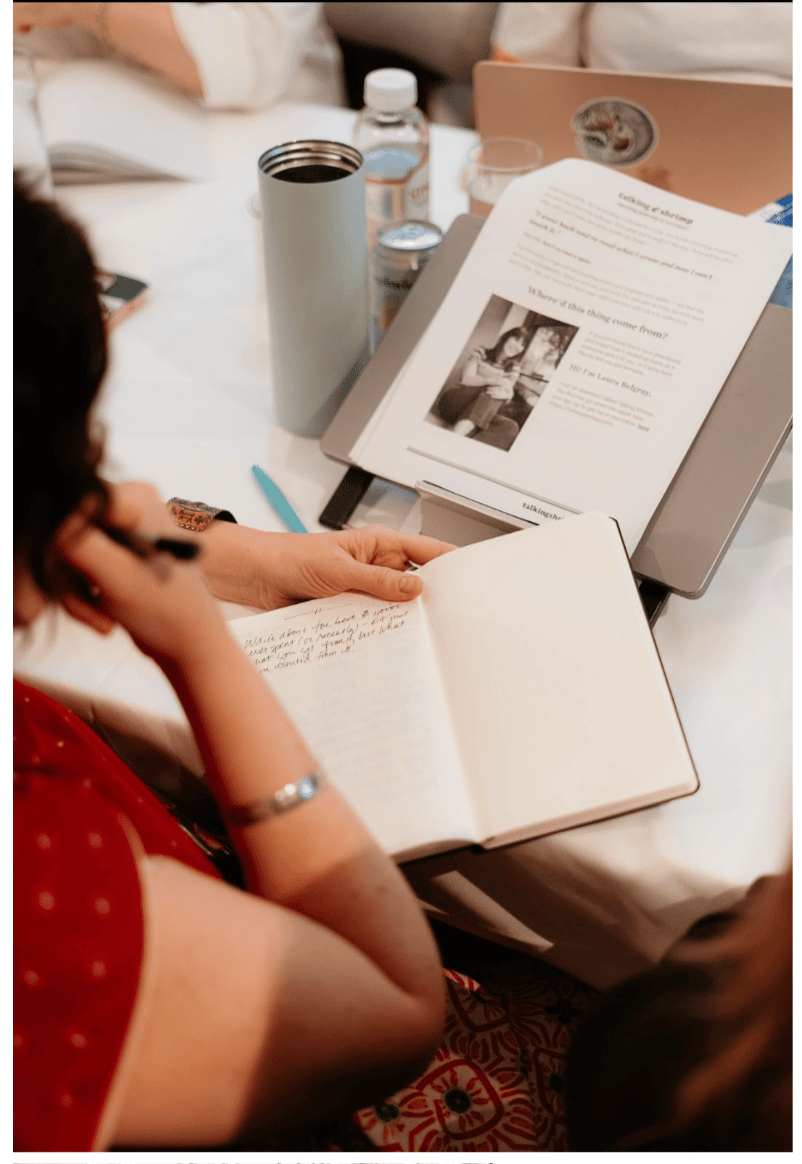 Person seated at a table writing in a notebook during a workshop, with a printed handout on a stand in front of them and a water bottle, tumbler, and laptop nearby.