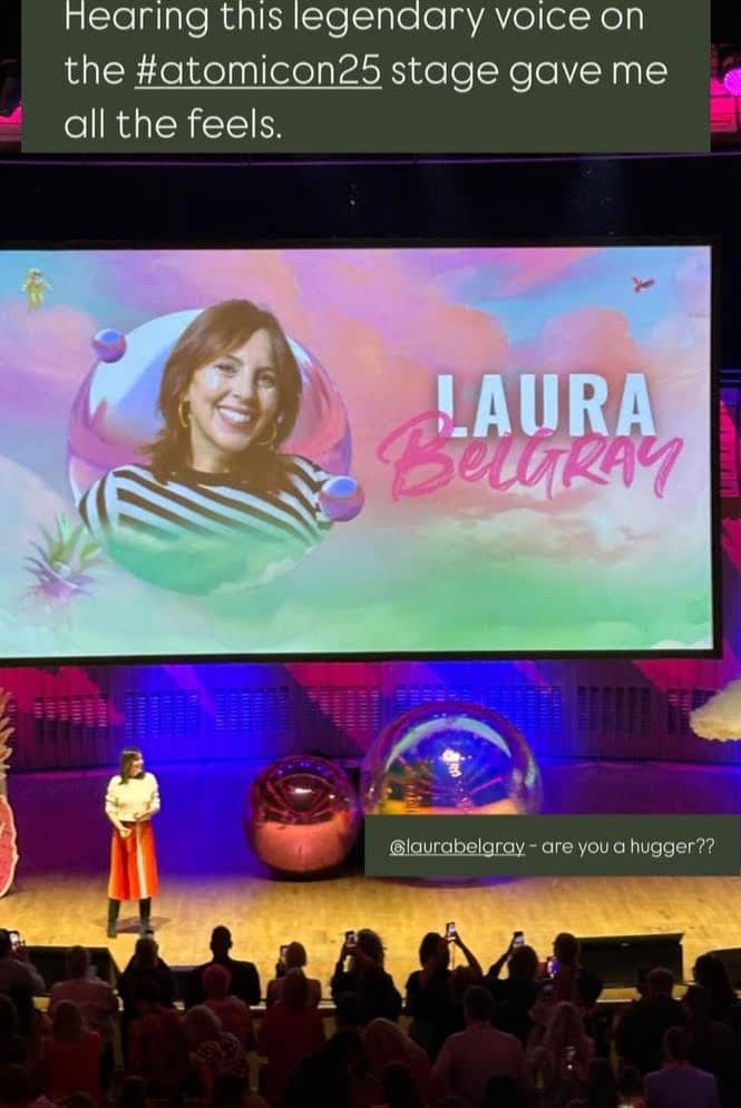 A photo from the Atomicon conference shows Laura Belgray speaking onstage in front of a large audience. Behind her, a massive screen displays her photo and name, “Laura Belgray,” against a colorful pastel background. Text overlaid on the image reads: “Hearing this legendary voice on the #atomicon25 stage gave me all the feels.” Another overlay near the bottom reads: “@laurabelgray - are you a hugger??” Audience members are standing and holding up their phones to take photos as Laura presents onstage.