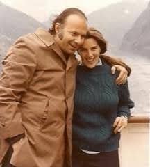 stand close together outdoors, smiling with their arms around each other. The man wears a tan jacket and the woman a dark blue sweater, with hazy mountains and water in the background.