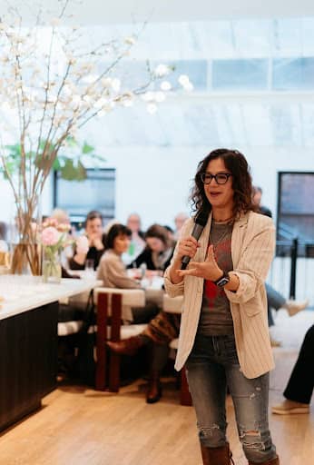 Woman with curly hair and glasses speaking into a handheld microphone while standing at the front of a bright workshop space, gesturing as attendees sit at tables behind her listening.
