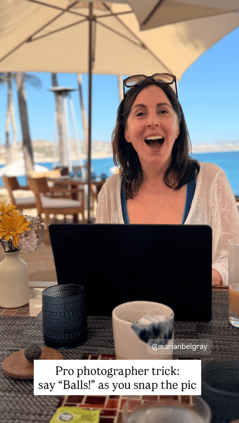Laura sits at an outdoor seaside café table with a laptop open in front of her, smiling widely with her mouth open as if mid-laugh. She has sunglasses resting on her head and sits under large patio umbrellas with the ocean and palm trees in the background. On the table are a small vase of flowers, cups, and drinks. Overlaid text reads: “Pro photographer trick: say ‘Balls!’ as you snap the pic.”