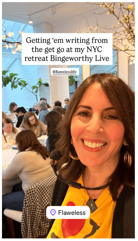 Laura Belgray in a bright yellow top taking a selfie inside a light-filled workshop space, while a group of people sit at tables behind her writing; on-screen text reads “Getting ‘em writing from the get go at my NYC retreat Bingeworthy Live” with a location tag “Flaweless.”