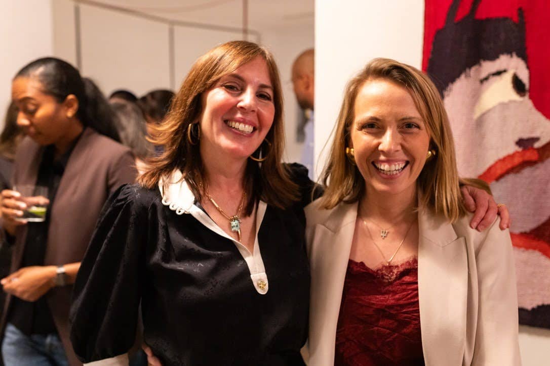 Laura Belgray and Candice stand together smiling at the Shrimp Club retreat venue, with Laura’s arm around Candice. Laura wears a black blouse with white trim and gold hoop earrings, and Candice wears a burgundy lace top with a white blazer. Other attendees and colorful artwork appear in the background.