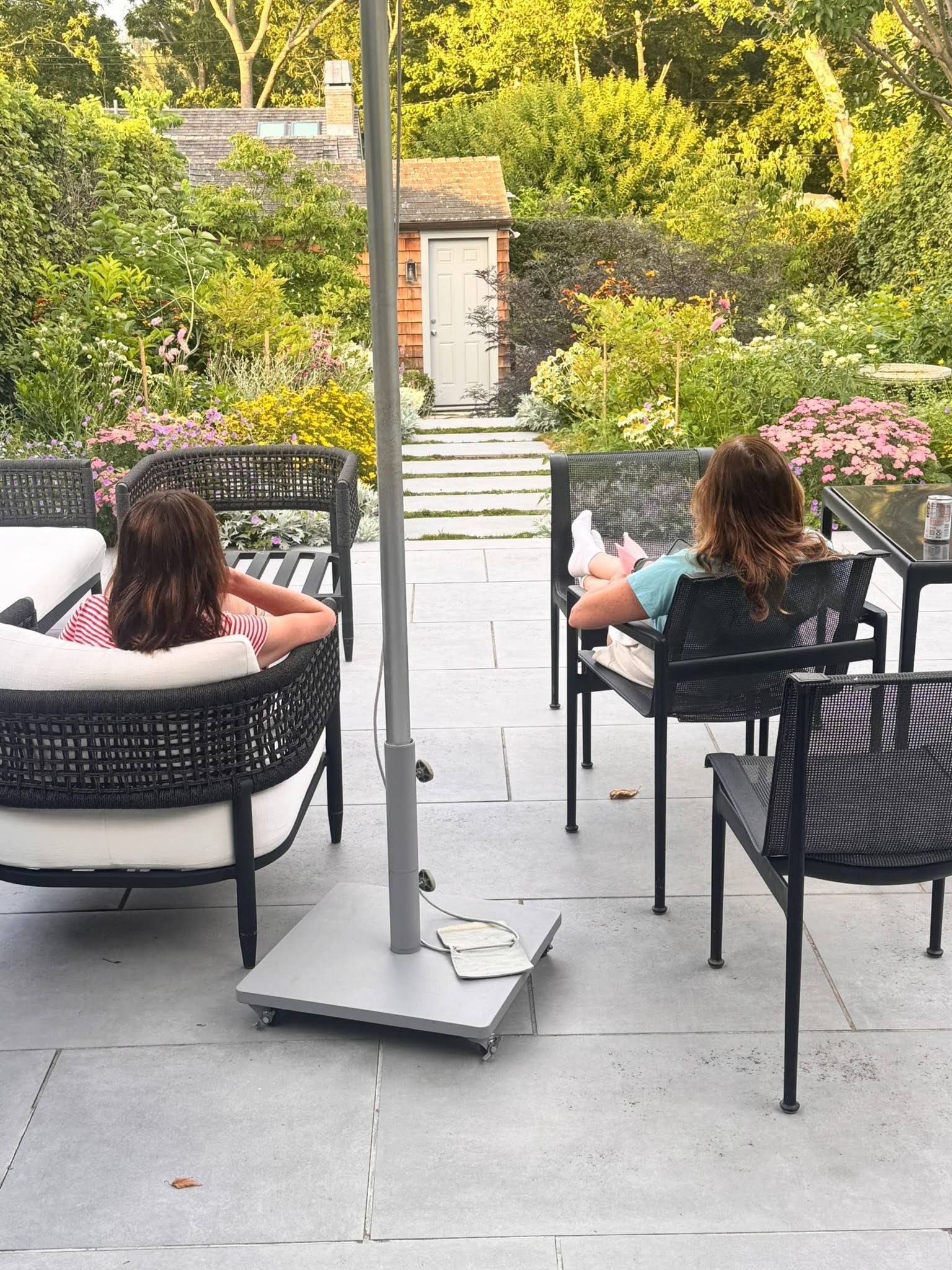 A photograph shows Laura Belgray and her sister relaxing on a spacious stone patio overlooking a lush, colorful garden. One has shoulder-length dark brown hair and is seated in a cushioned woven lounge chair wearing a red-and-white striped shirt. The other has medium-length brown hair with lighter ends and is reclining in a black mesh chair with her feet up, reading a book. A large patio umbrella stands in the center of the patio. Flower beds filled with pink, yellow, white, and purple blooms line a stone walkway leading to a small garden shed with a white door. Mature trees and dense greenery surround the peaceful backyard, and a can of sparkling water sits on the outdoor dining table to the right.