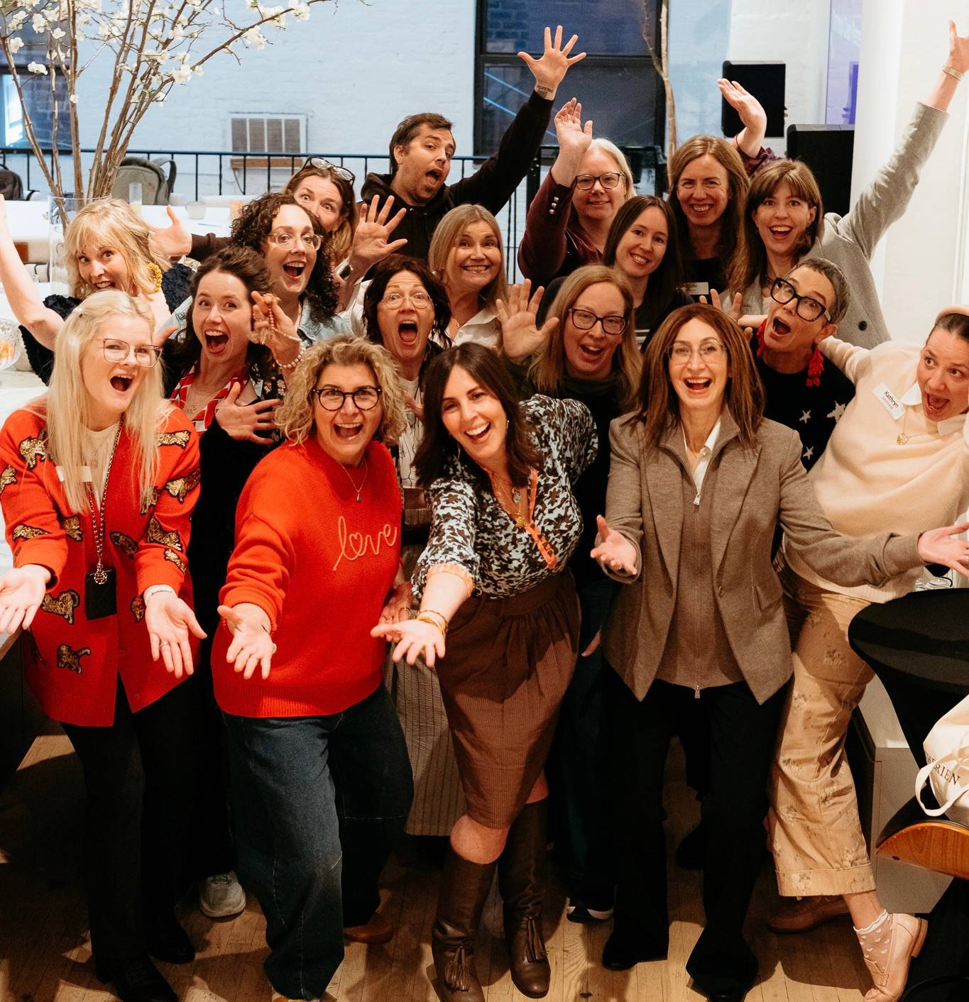 A group of about 20 smiling adults strikes playful poses with raised arms and open hands in a bright loft space.
