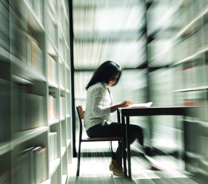 A woman sitting at a table in a library, writing in a notebook, surrounded by tall bookshelves with a soft motion blur effect suggesting focus and momentum.