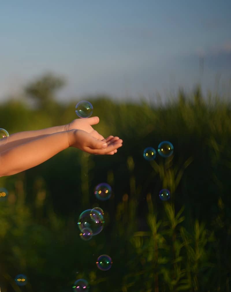 Hands reaching for iridescent soap bubbles in a green field.