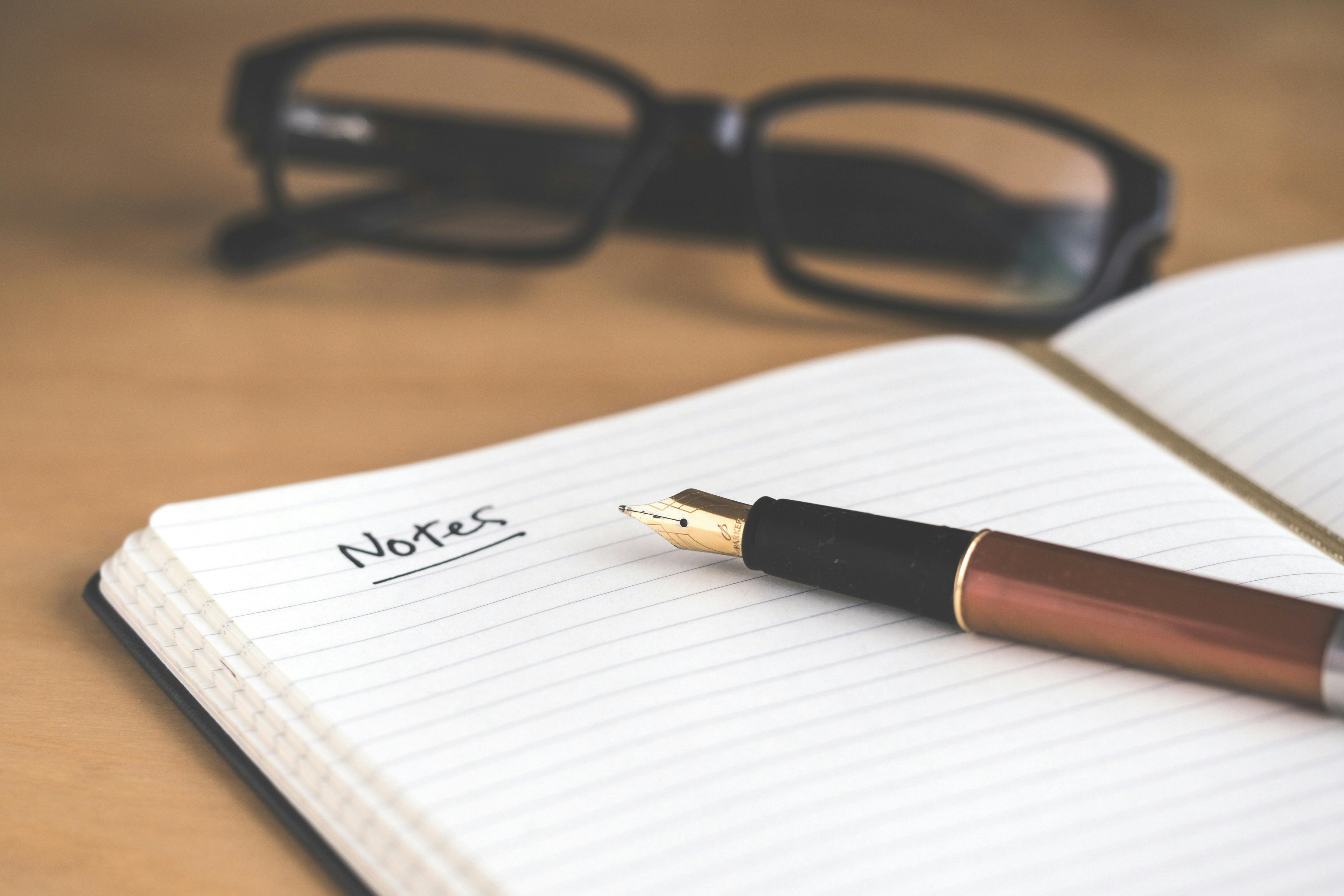 fountain pen on top of an open notebook, and black rimmed glasses on background all on a light brown desk