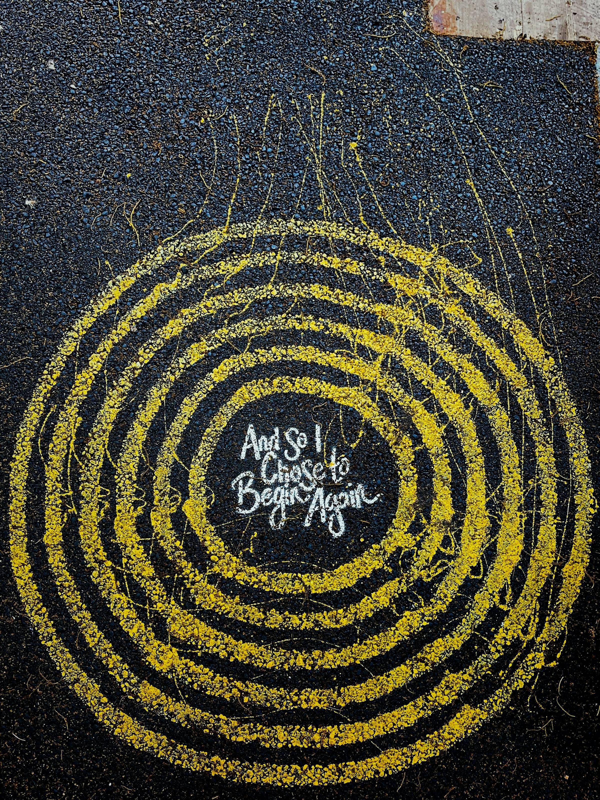 yellow concentric circles on concrete that reads "and so i choose to begin again" at the center