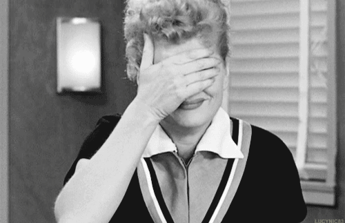 Lucille Ball in black-and-white with her hand covering her face. She peeks through her fingers with one eye.