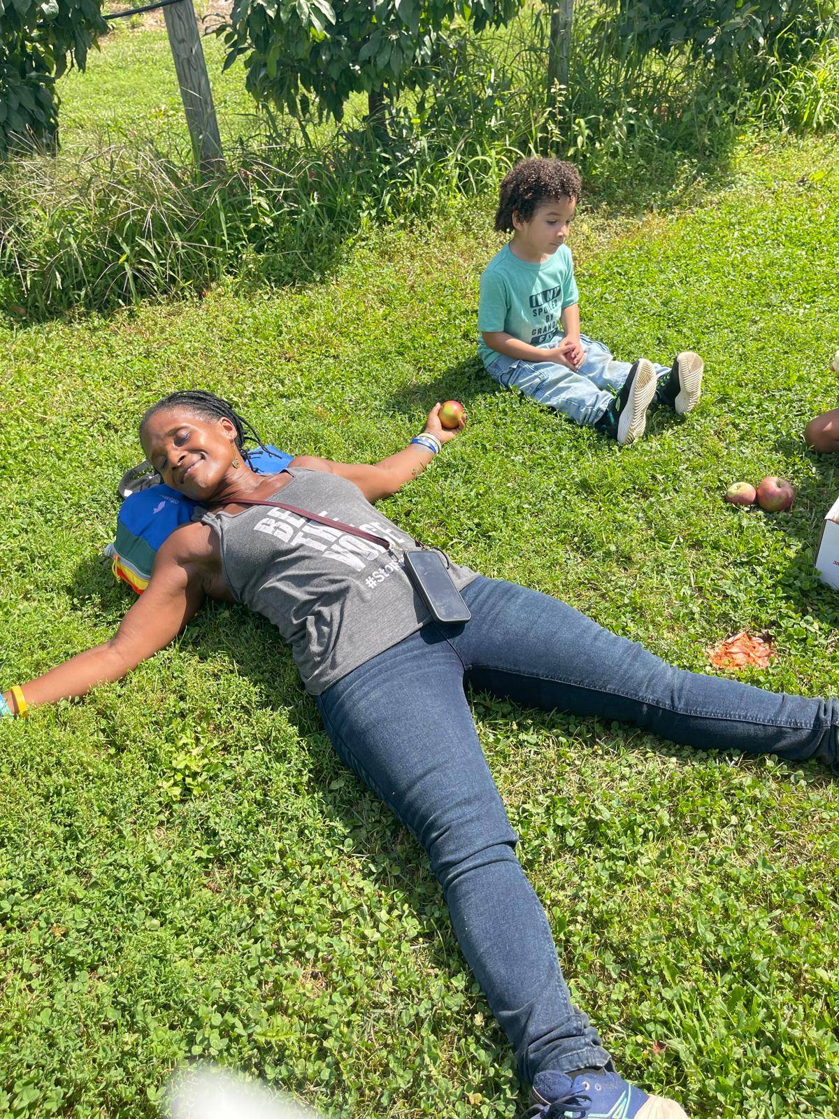 Dionne lying in the grass holding an apple while her grandson sits nearby, both surrounded by scattered apples in an orchard, relaxed and peaceful in the sun.
