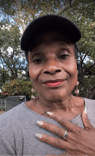 A candid outdoor photo of Dionne wearing a cap and resting a hand on her chest, surrounded by fall trees. The image reflects a moment of presence, grounding, and personal expression.