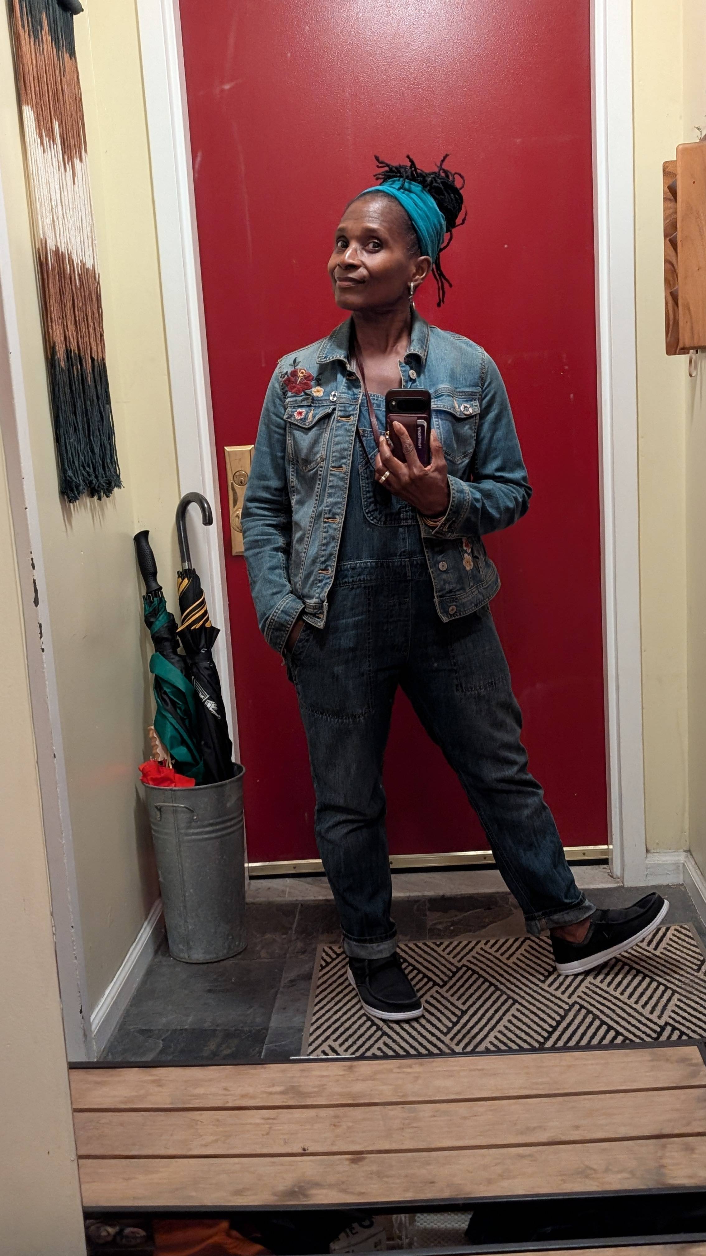 A mirror selfie of Dionne standing in her entryway wearing denim overalls, a teal headwrap, and a floral?embroidered jacket. She looks grounded and calm, capturing the moment she returned home on the day vertigo began — a reminder that people don’t always
