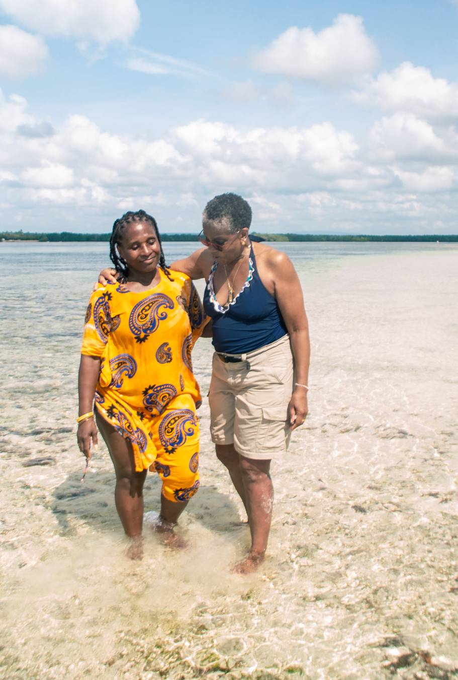 Two women walking together in shallow beach water, one wearing a bright yellow dera and the other in navy and beige. They move forward with no bags, capturing companionship, ease, and the spiritual spaciousness of traveling light.