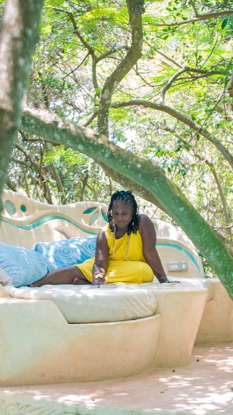 A woman in a bright yellow dress sits on an outdoor bed surrounded by lush green trees, journaling in soft natural light. Blue patterned pillows and an artistic sculpted headboard frame the scene, creating a peaceful, reflective atmosphere.