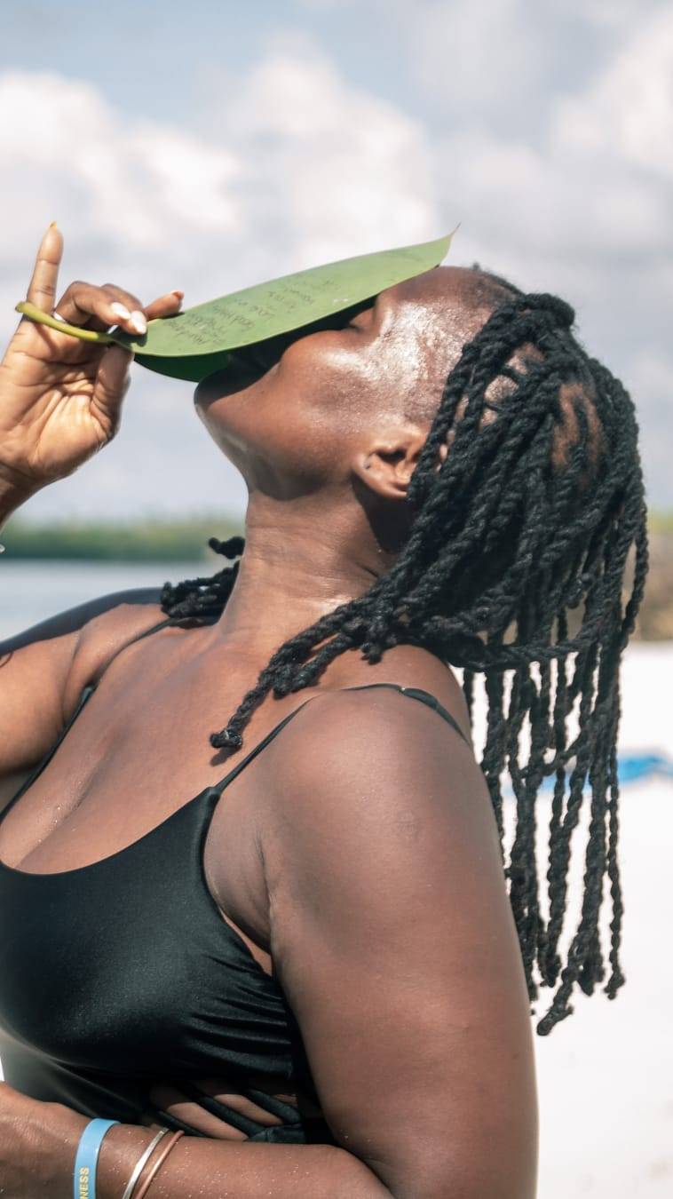 A woman holds a large green leaf near her face while looking upward toward the sky. Her hair is styled in twists, and she stands outdoors with soft clouds and a blurred shoreline behind her, creating a peaceful, contemplative scene.