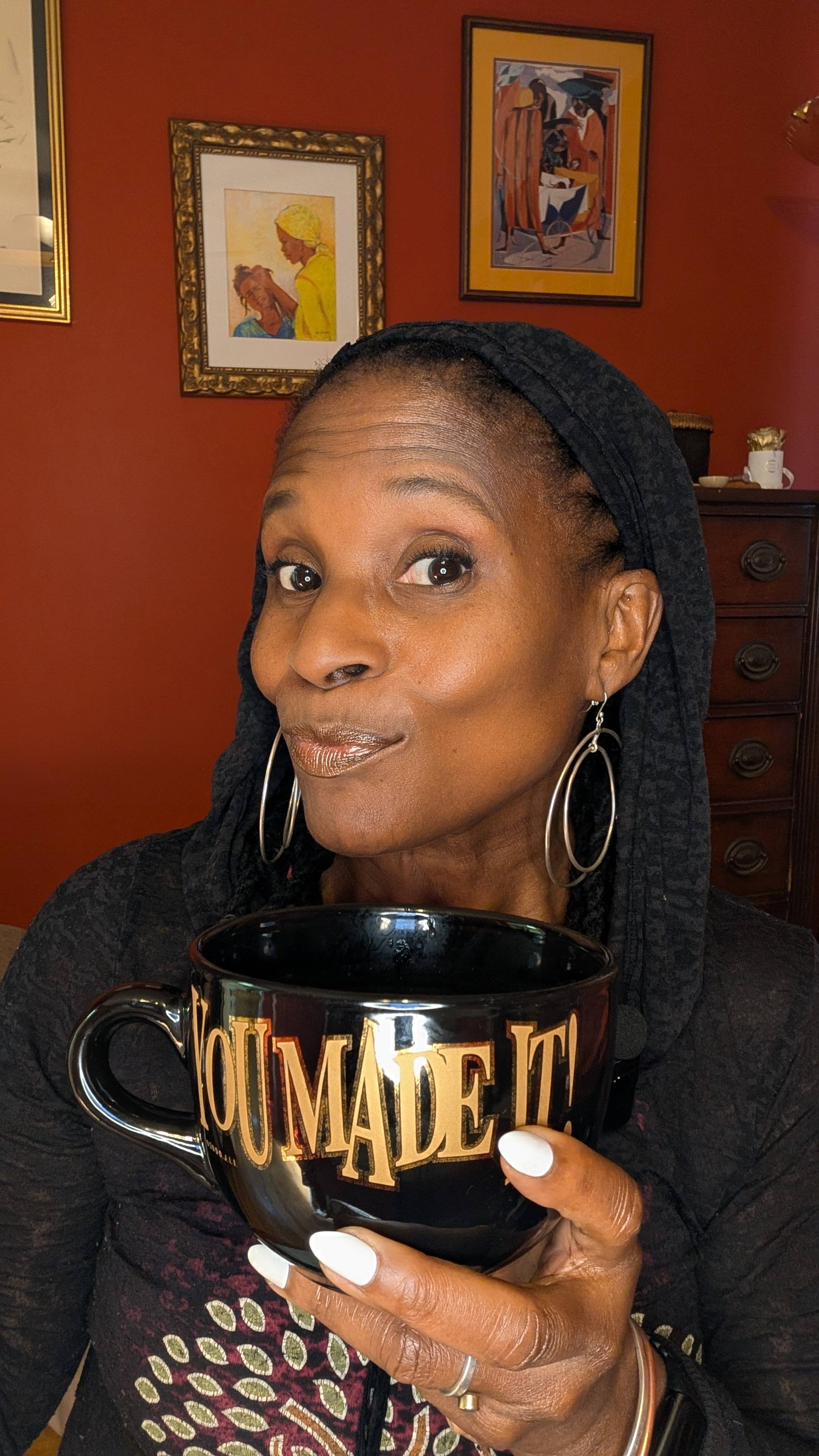 A close-up of Dionne holding a black mug that says “YOU MADE IT!” in gold lettering, sitting in her red-walled home with artwork behind her — a calm, grounded moment before the story unfolds.