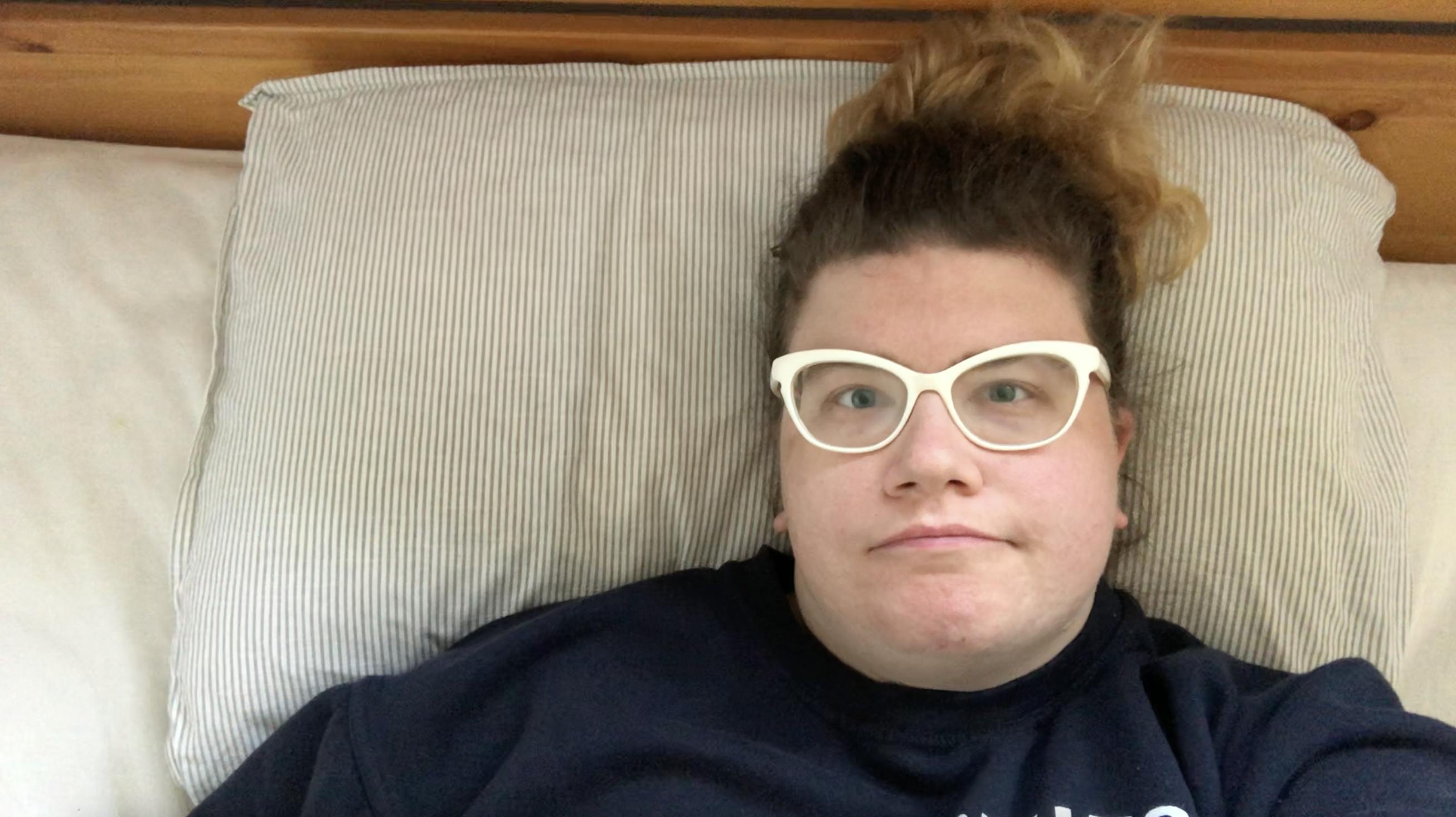 white woman with white glasses and blonde hair wearing a blue sweatshirt lying on her back in bed on a striped pillow and wooden bedframe