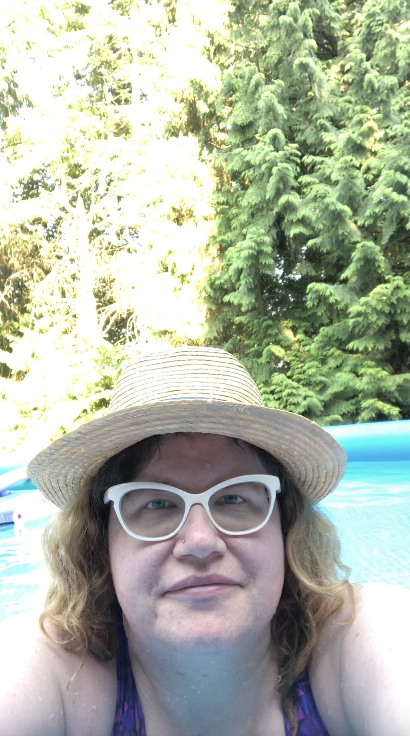 white woman with blonde curly hair wearing white frame glasses and a straw hat and purple swimsuit in a blue pool in front of green cedar trees