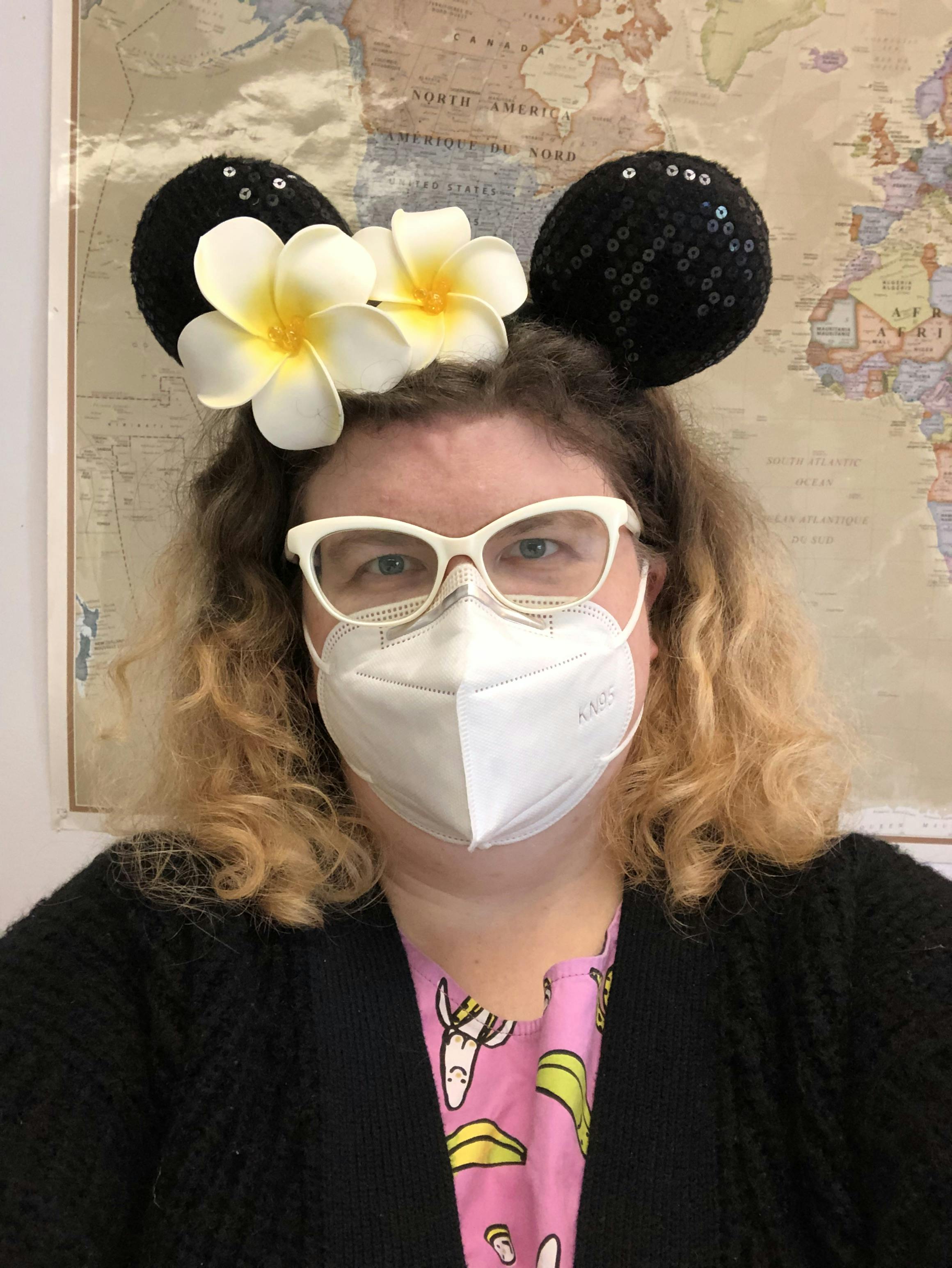 white woman with curly hair and white framed glasses wearing a white face mask and black sequined Disney mouse ears with white flowers