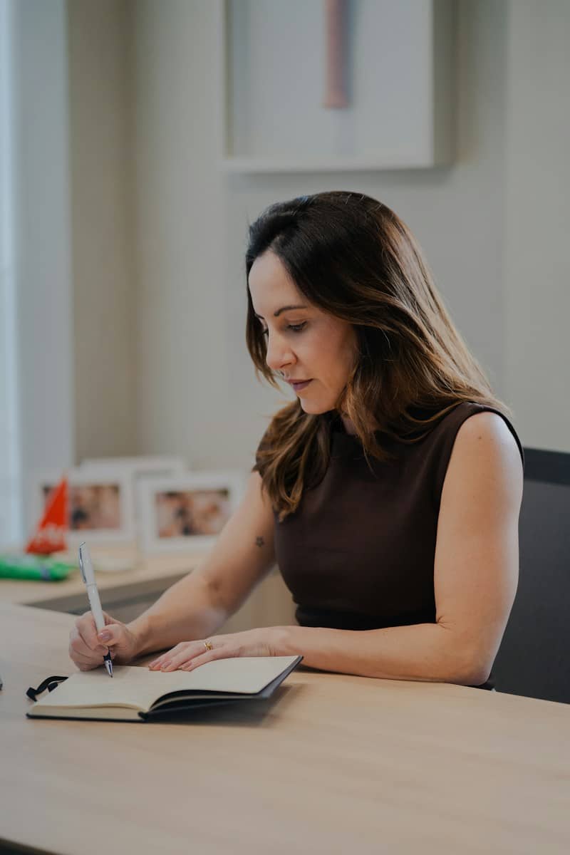 A woman in a brown sleeveless top writing in a notebook at a desk