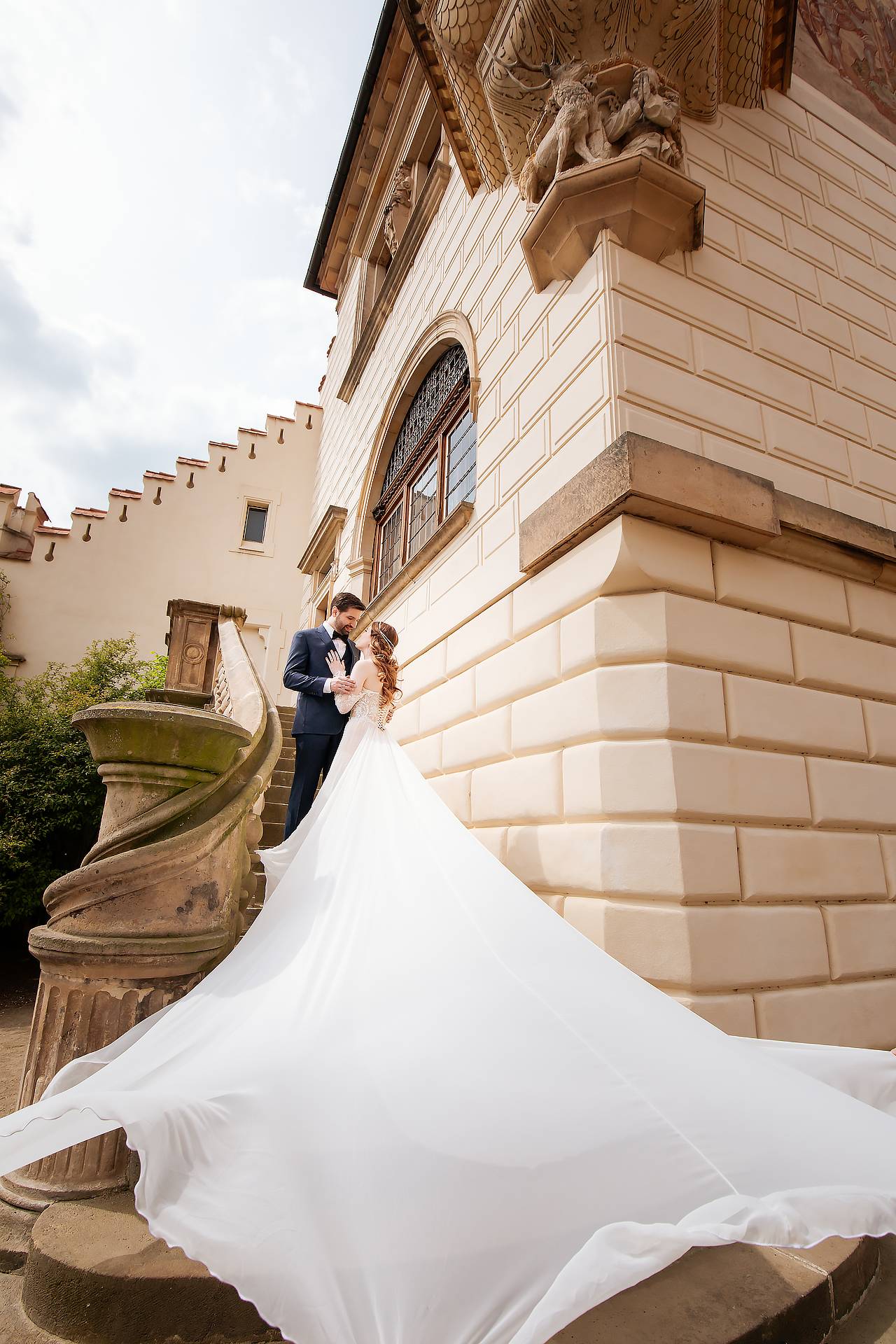 Me in a ridiculously long, flowy wedding dress standing on the steps of Pruhonice Castle in Prague embracing my husband