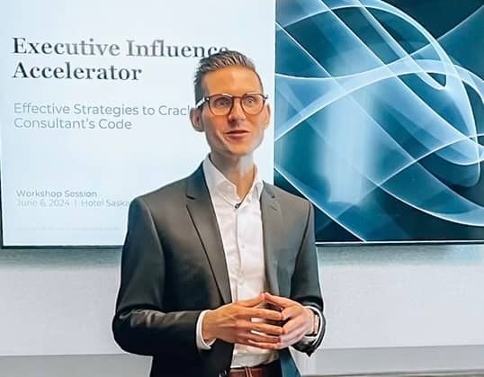 Photo of Brennen Schmidt wearing a black suit jacket and glasses standing in front of a screen with the title "Executive Influence Accelerator"